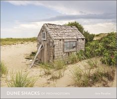 an old shack sitting on top of a sandy beach next to tall grass and bushes