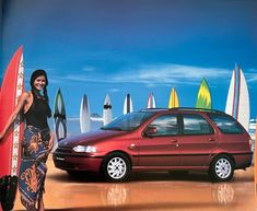 a woman standing next to a red car with surfboards on the beach behind her