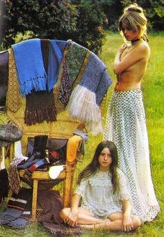 two women sitting in the grass next to an old chair with clothes hanging on it