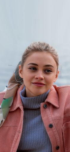 a woman wearing a pink jacket and grey sweater is posing for the camera with her hair in a bun