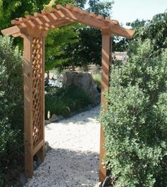 a wooden pergoline in the middle of a gravel path surrounded by bushes and trees