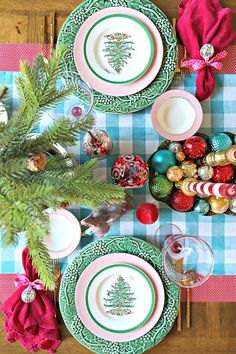 the table is set for christmas dinner with green and pink plates, silverware, red napkins, pine tree ornament