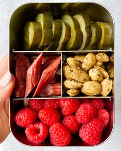 a person is holding a lunch box filled with fruits and veggies, including pickles