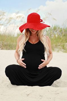 a pregnant woman sitting on the beach wearing a red hat and black dress with her hands in her stomach