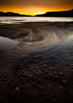 the sun is setting over an ocean with sand and mountains in the background, as seen from across the water