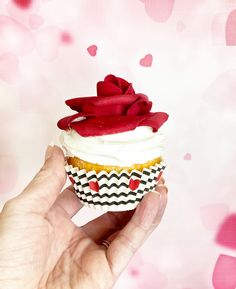 a hand holding a cupcake with white frosting and a red rose on top