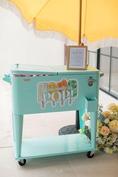 an old fashioned ice cream machine with flowers on the side and a yellow umbrella over it