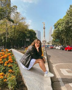 a woman sitting on the edge of a wall with flowers in front of her and buildings behind her