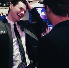 a man in a suit and tie smiles at another man who is adjusting his hat