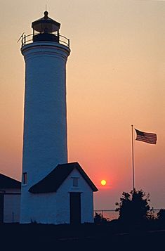 a lighthouse with the sun setting behind it and an american flag flying in front of it