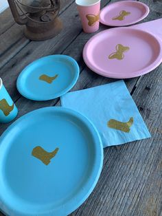 pink and blue plates with gold designs on them sitting on a wooden table next to cups