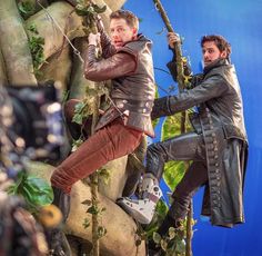 two men in leather jackets climbing up a tree with camera equipment on the ground behind them