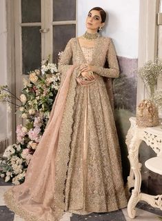 a woman standing in front of a wall wearing a beige and gold lehenga