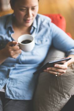 a woman sitting on a couch holding a cup of coffee and looking at her cell phone