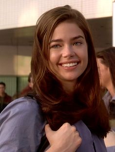 a woman with long brown hair and blue shirt smiling at the camera while standing in front of a crowd