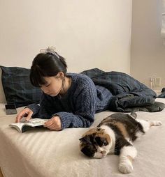 a woman laying on top of a bed reading a book next to a calico cat