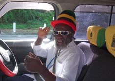 a man sitting in the passenger seat of a car wearing a hat and sunglasses, holding his hand up