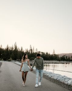 a man and woman holding hands while walking down the road next to a body of water