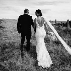 a bride and groom walking through the field holding hands with their veil blowing in the wind