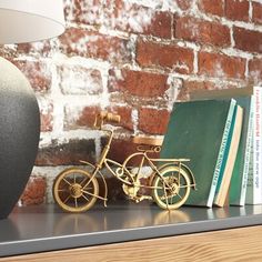 there is a small gold bike next to a book and lamp on a shelf in front of a brick wall