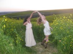 two women in white dresses are dancing on a path through tall grass and wildflowers