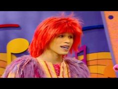 a man with red hair and pink wig standing in front of a blue background wearing a purple outfit