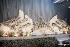 a stage set up for a wedding with white chairs and flowers on the wall behind it