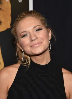 a woman with blonde hair and big hoop earrings smiles at the camera while standing in front of a movie poster