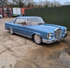 an old blue car parked in front of a green fence and some shipping containers behind it