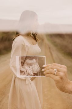 a woman is holding up a photo in front of a man's face on a dirt road