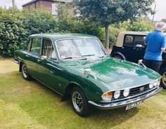 an old green car is parked in the grass with people looking at it from behind