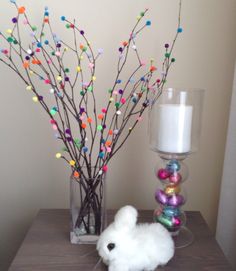 a small white bunny sitting next to a vase filled with easter eggs and branches in front of a candle