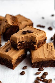 several pieces of chocolate brownie with frosting and coffee beans scattered around it on a white surface