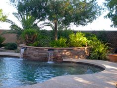 a pool with a waterfall in it next to a tree and shrubbery on the other side