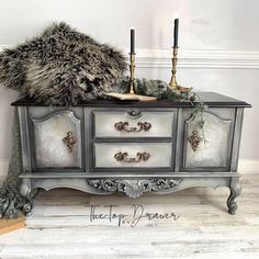 an ornate dresser with two candles and a fur pillow on it's top, in front of a white wall