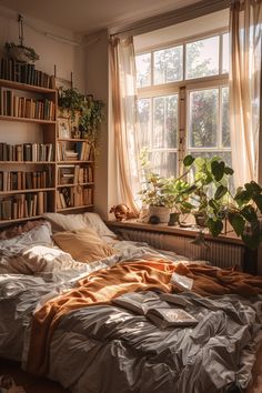 an unmade bed sitting in front of a window with bookshelves and plants