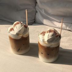 two drinks with whipped cream and cinnamon sticks in them sitting on a white tablecloth