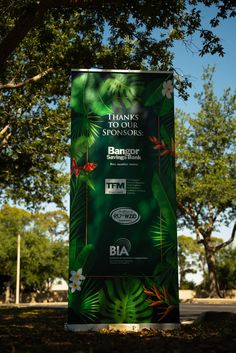 a large green sign sitting in the middle of a park next to a lush green tree