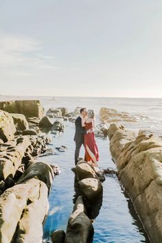 a man and woman standing on rocks near the ocean with their arms around each other