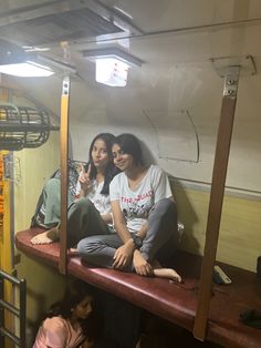 two women sitting on top of a bunk bed in a room with other people around