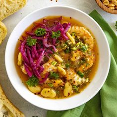 a white bowl filled with shrimp and red cabbage soup next to sliced bread on a table