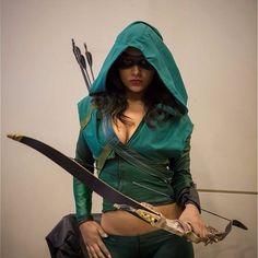 a woman dressed in green holding two arrows and wearing a hoodie with her hands on her hips