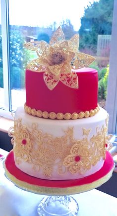 a three tiered cake with red and gold decorations on the top, sitting in front of a window