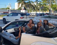 three women sitting in the back of a convertible car with their hands up and two men standing next to them