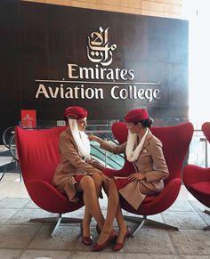 two women sitting on red chairs in front of emirates's aviation college sign