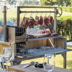 meat is being cooked on an outdoor grill with wine glasses and utensils next to it