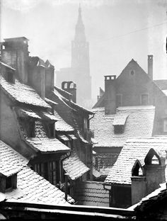 an old black and white photo of rooftops with snow on the roofs, and a clock tower in the background