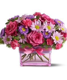 pink and purple flowers in a square glass vase with ribbon around the edges, on a white background