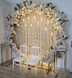 two white chairs sitting next to each other in front of a wall with flowers and lights