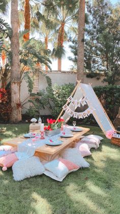 a picnic table set up in the grass with pink and white pillows, plates and utensils